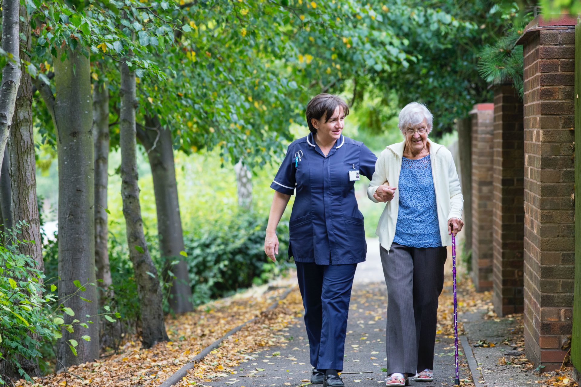 Older lady with walking stick, walking towards the camera supported by female NHS employee