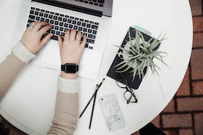 Aerial view of desk with hands over a laptop keyboard, pot plant, glasses and pen